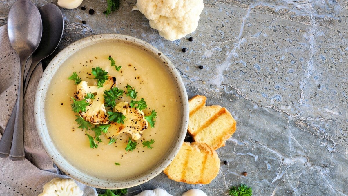 Eine graue Schale mit der hellgelben Suppe auf einem hellen Marmoruntergrund. Daneben Löffel, Knoblauch und geröstetes Brot.