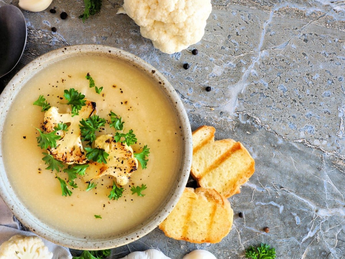 Eine graue Schale mit der hellgelben Suppe auf einem hellen Marmoruntergrund. Daneben Löffel, Knoblauch und geröstetes Brot.