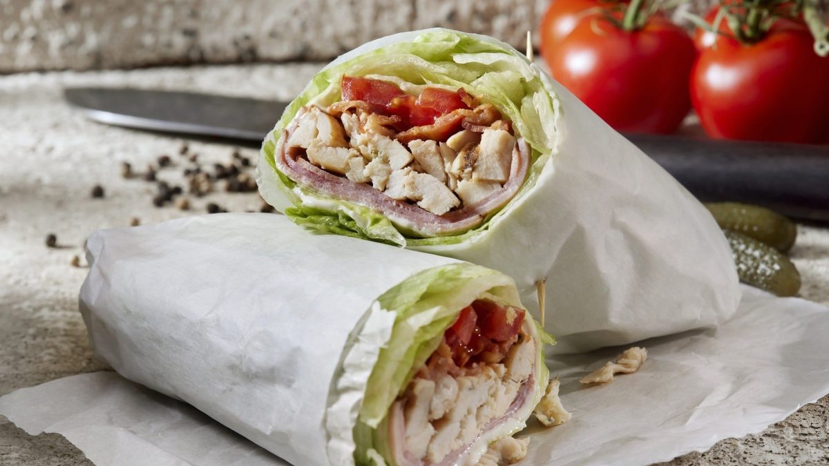 Brathähnchen-Wrap mit Salat, Tomate, Schinken und Bacon, angeschnitten serviert auf einem Backpapier.