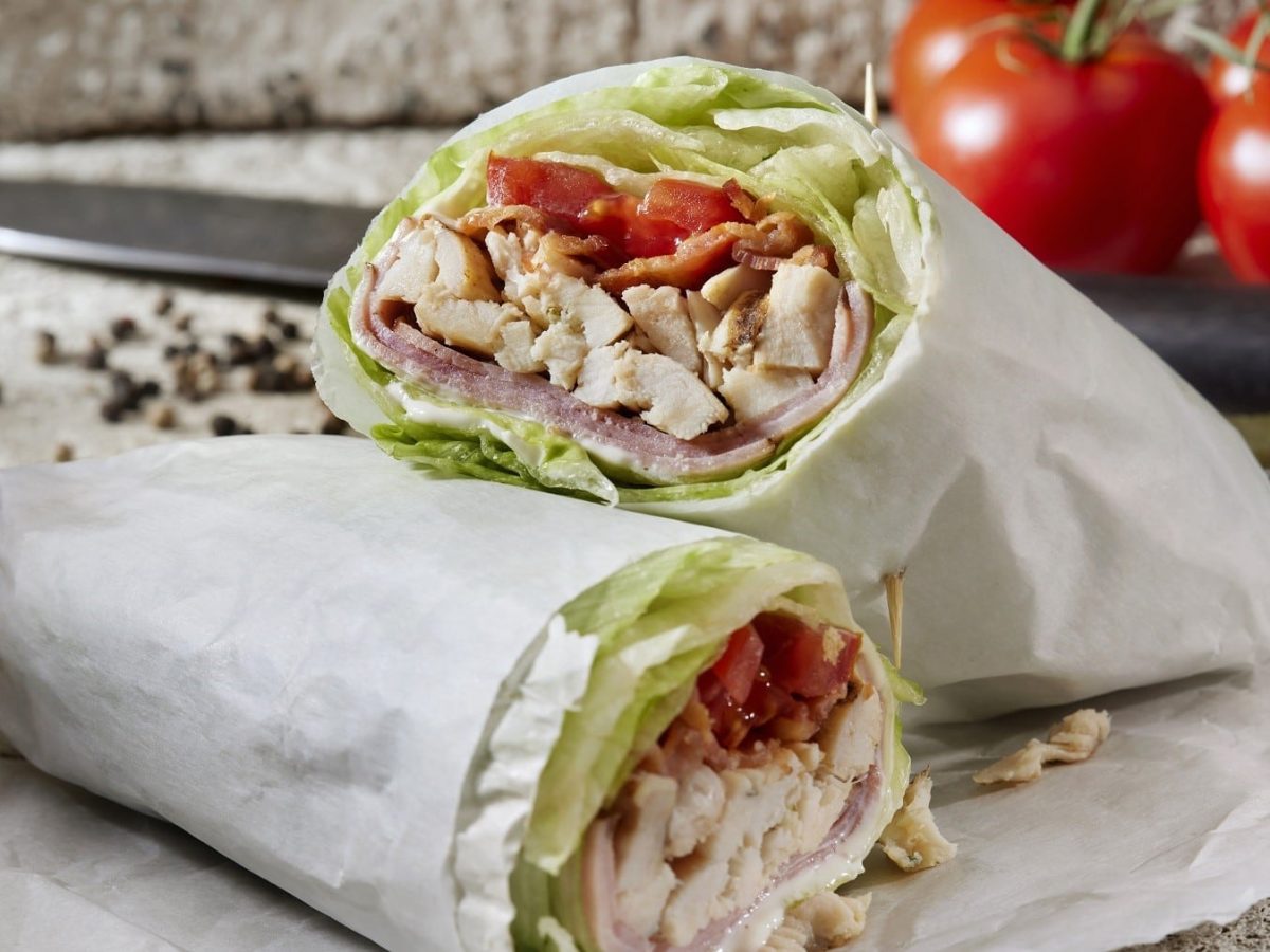 Brathähnchen-Wrap mit Salat, Tomate, Schinken und Bacon, angeschnitten serviert auf einem Backpapier.