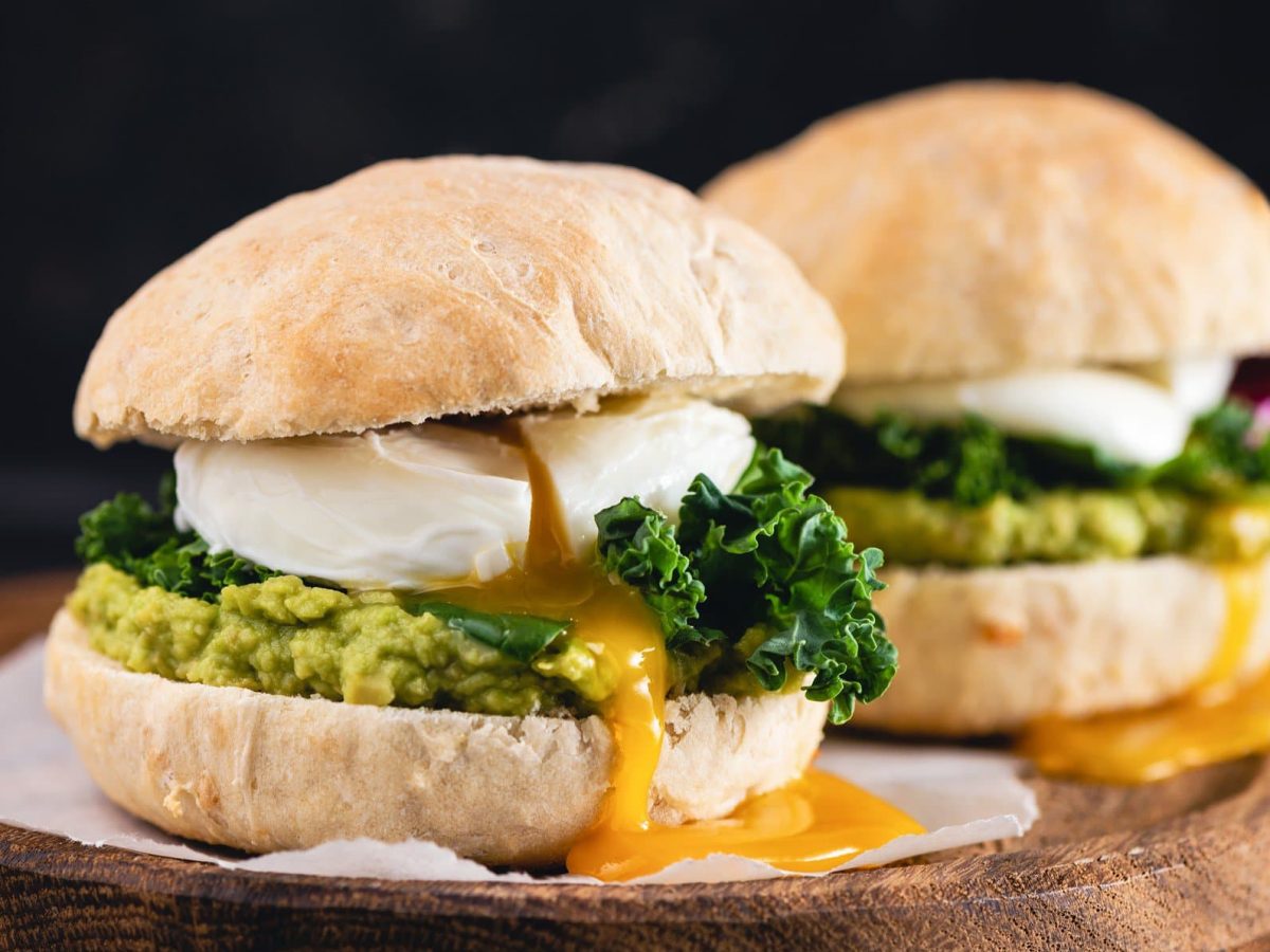 Breakfast-Burger mit pochiertem Ei, Schwarzkohl und Avocadosalsa im Brioche auf Krepp und dunklem Holz. Frontalansicht.