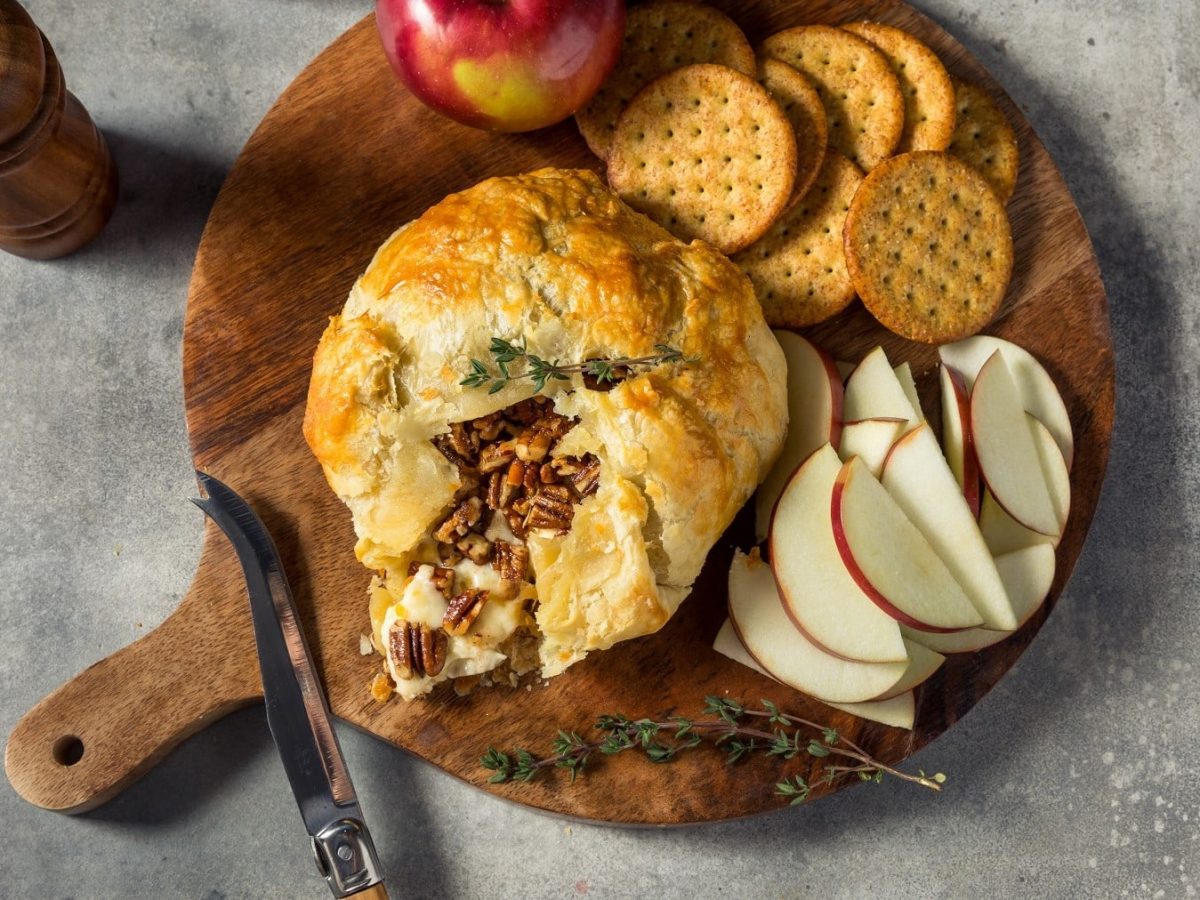 Brie-Pie mit Apfel und Pekannüssen gefüllt, angeschnitten serviert auf einem Holzbrett und angereicht mit Crackern und frischen Apfelscheiben.