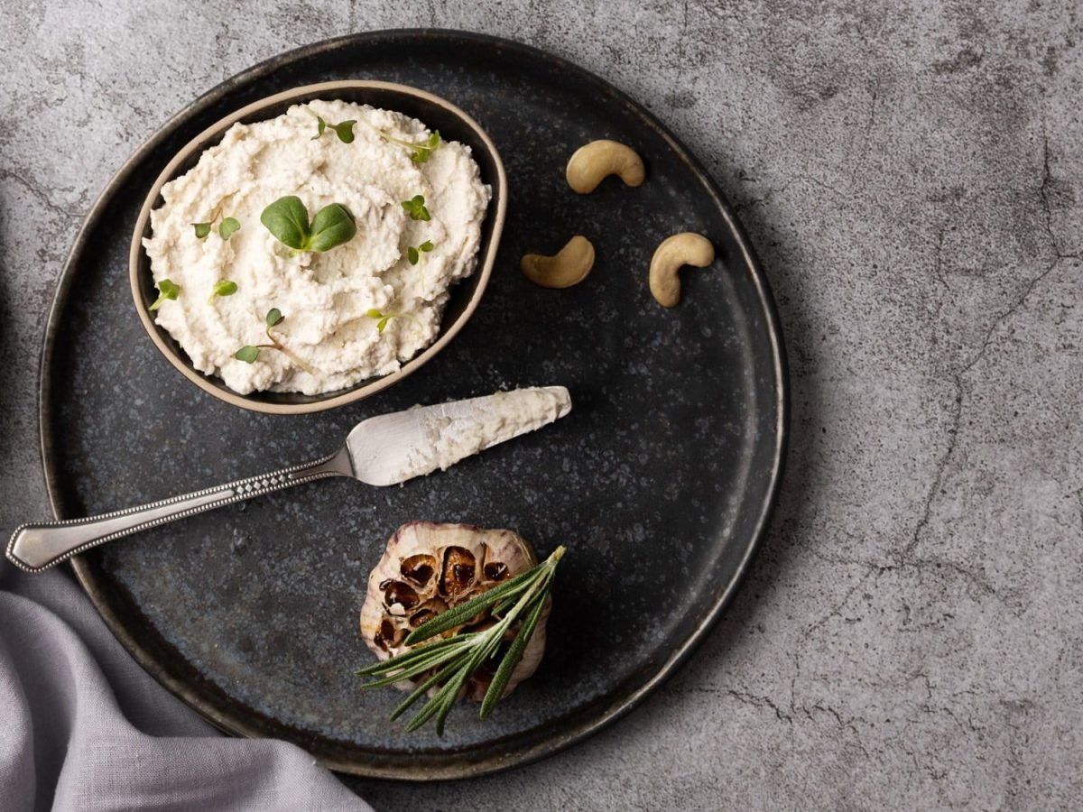 Eine Schüssel Cashew-Hummus auf einem Teller, darauf liegen außerdem Cashews und ein Messer sowie eine gebackene Knoblauchknolle. Draufsicht.