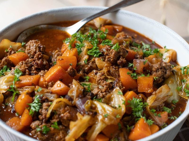 Eine weiße Schüssel mit dem Chinakohl-Eintopf auf einem dunklen Holztisch. Oben rechts ist ein silberner Löffel im Eintopf.