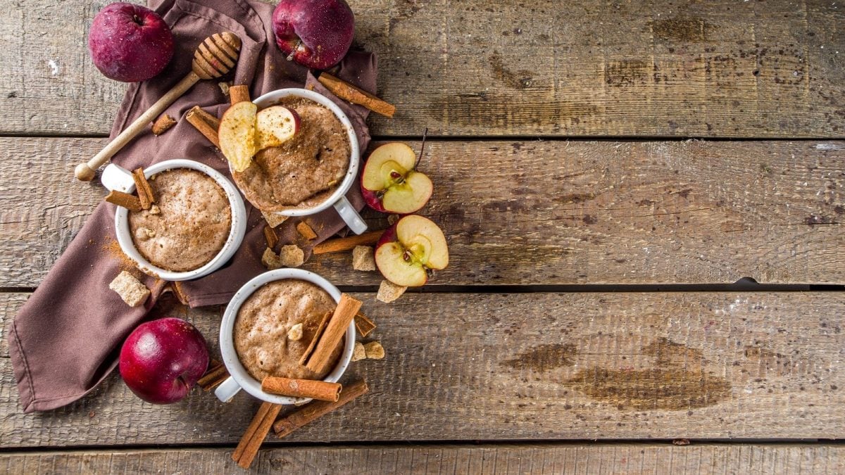 Drei Apfel-Zimt Mug Cakes neben Äpfeln und Zimtstangen auf einem Holztisch.