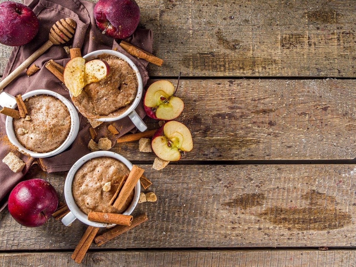 Drei Apfel-Zimt Mug Cakes neben Äpfeln und Zimtstangen auf einem Holztisch.