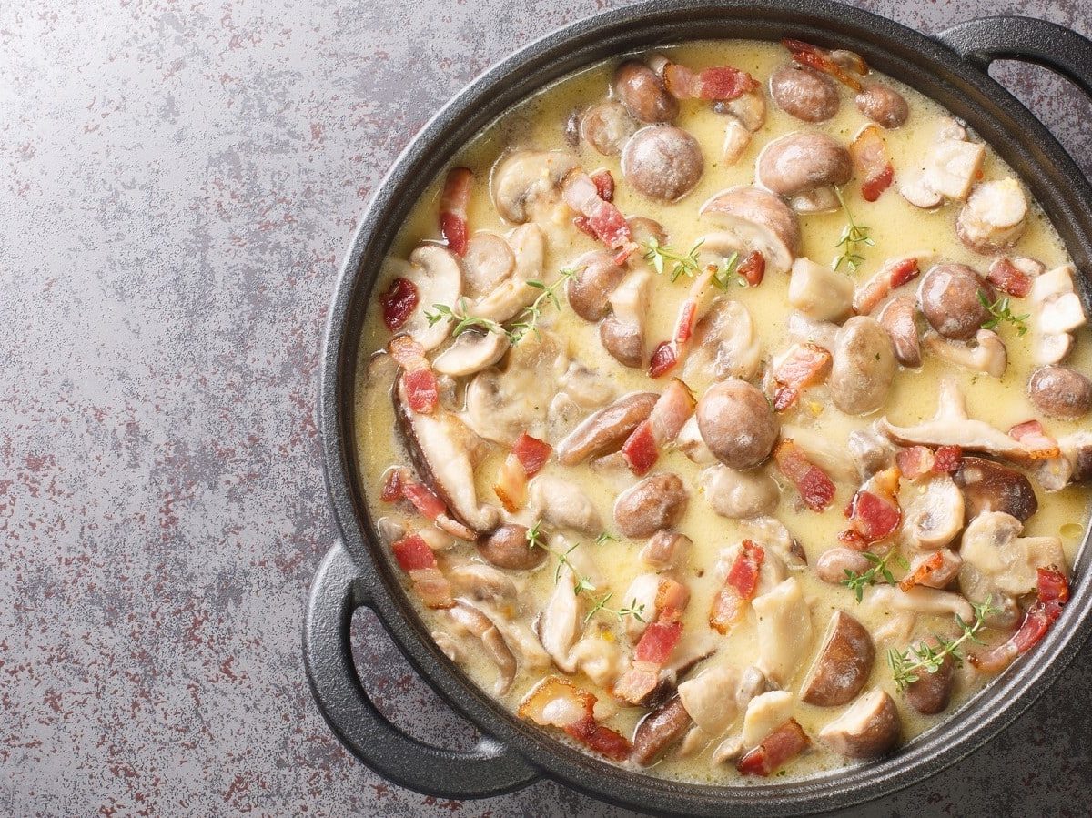 Ein schwarzer Topf mit Champignon-Speck-Sauce auf einem grauen Tisch.