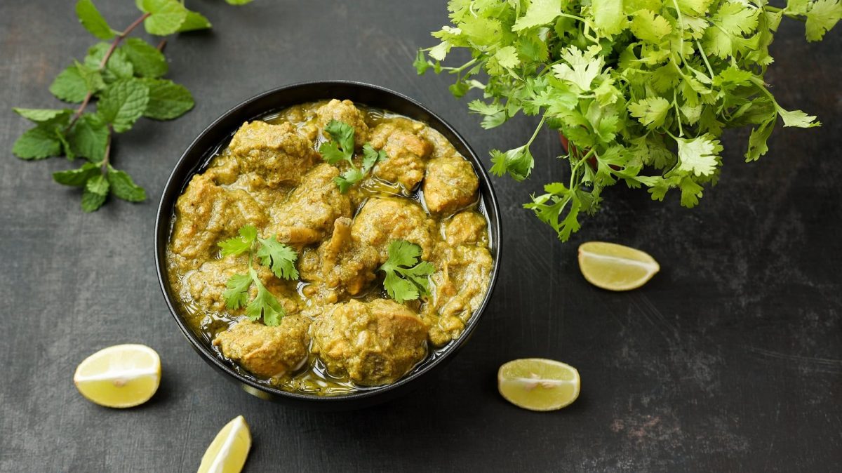 Eine schwarze Schüssel mit Hariyali Chicken Tikka auf einem schwarzen Untergrund neben Limettenspalten.