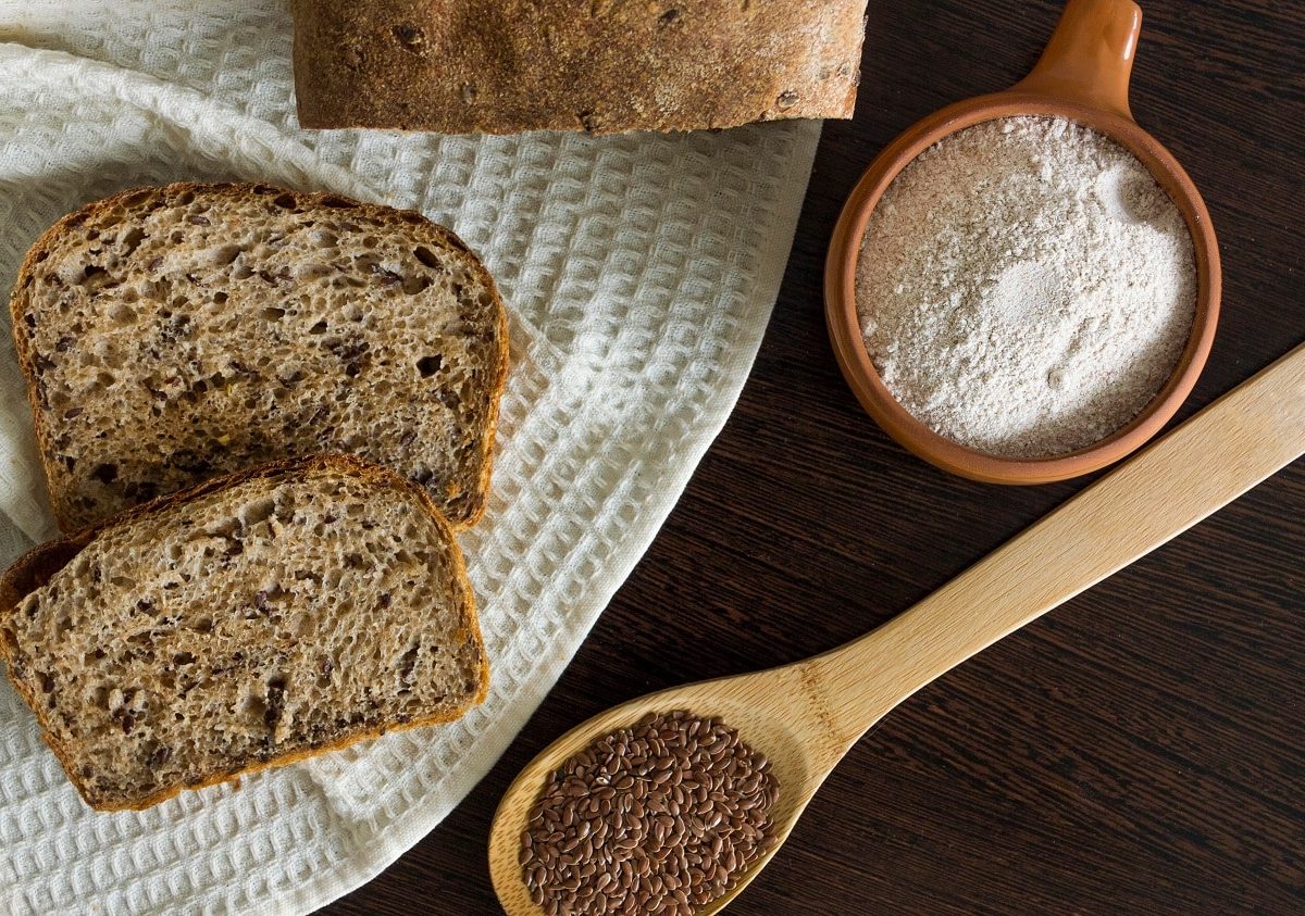 Draufsicht: Auf einem dunklen Untergrund ist ein kleiner Teil eines Leinsamenbrotes zu sehen. Daneben liegen zwei Scheiben Brot, ein Holzlöffel mit leinsamen und eine Schüssel mit Mehl.