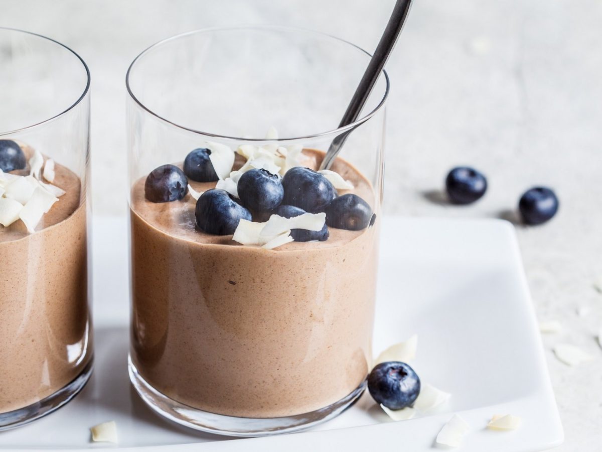 Zwei Gläser vegane Erdnussbutter-Mousse mit Blaubeeren und Kokosflocken, darin Teelöffel.