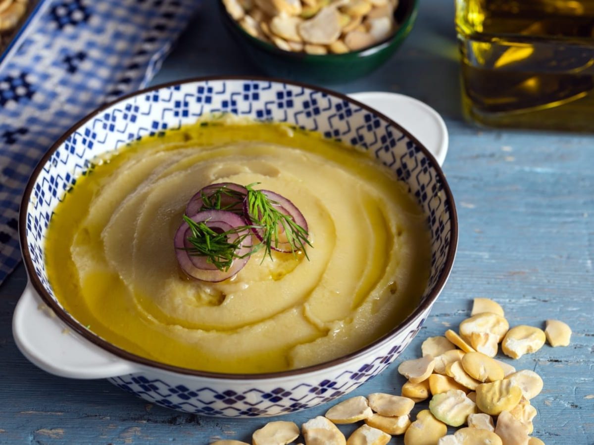 Blauer Untergrund mit einer Schüssel im Bildzentrum, welche mit einem griechisch-blau-weißem Muster bemalt ist. Darin ein cremiger Fava Aufstrich. Garniert mit frischer Petersilie. Um die Schüssel verteilt liegen lose Fava-Bohnen und frisches Olivenöl.