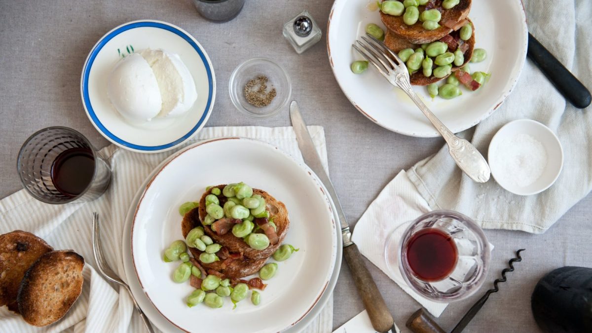 Draufsicht: Grauer Tisch angerichtet mit Stoffservierten und zwei weißen Tellern auf denen je zwei Scheiben Baguette mit Favabohnen und Speck liegen. Dazu auf einem weiteren Tellerchen eine Kugel frischen Mozzarella. Außerdem weitere Baguette-Scheiben und ein Glas Rotwein.