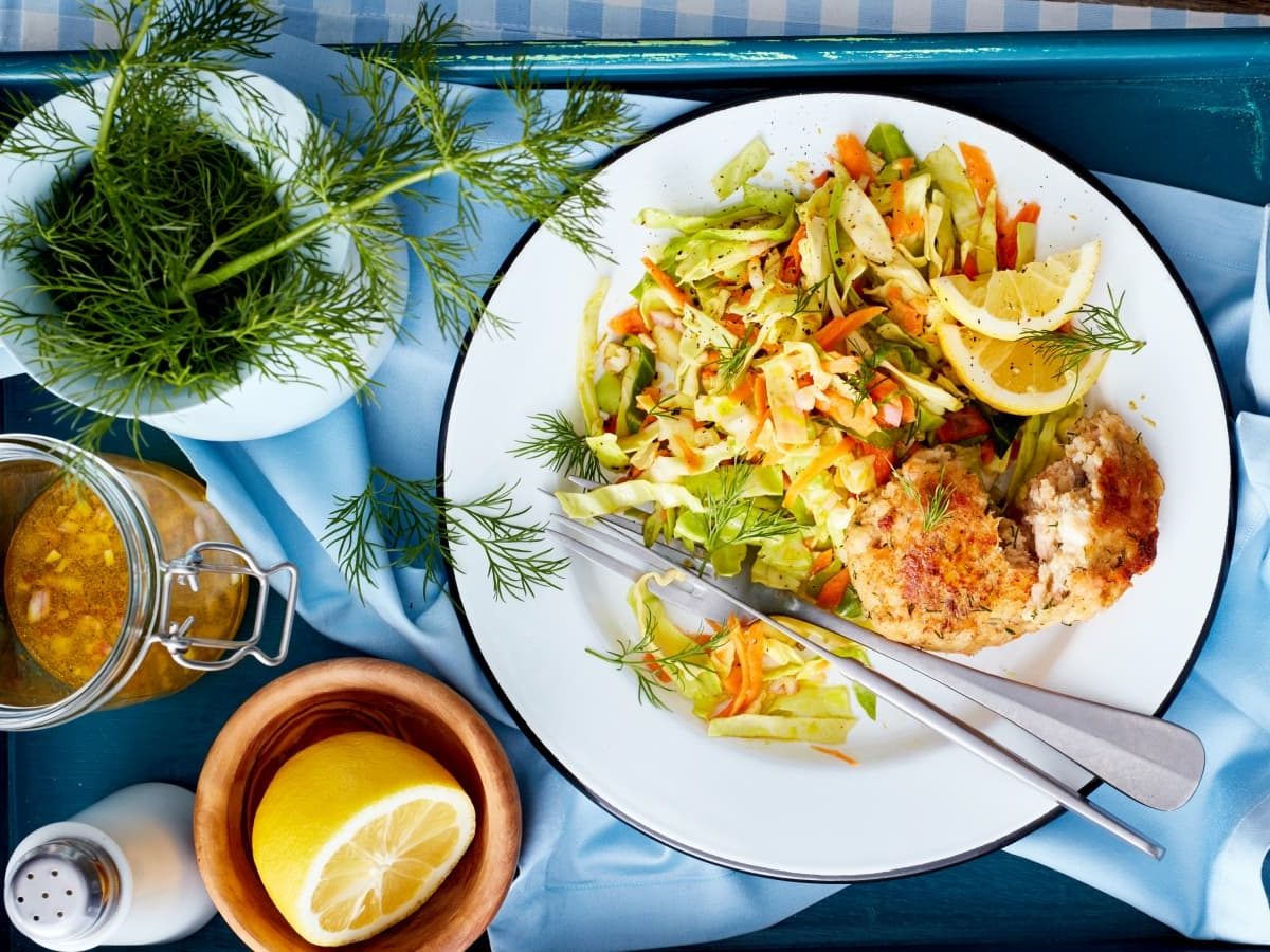 Draufsicht: Holztisch mit einem blauen Tablett auf dem auf der rechten Seite ein Teller mit Forellen-Frikadellen und Coleslaw steht. Auf der linken Seite ein Kännchen mit frischem Dill, ein Bügelglas mit Dressing und einer halben Zitrone, sowie einem Salzstreuer.