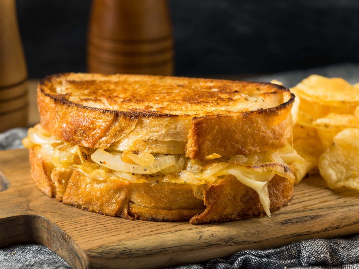 French Onion Cheese Sandwich mit Chips auf Holzbrett. Im Hintergrund Salzstreuer aus Holz. Frontalansicht.