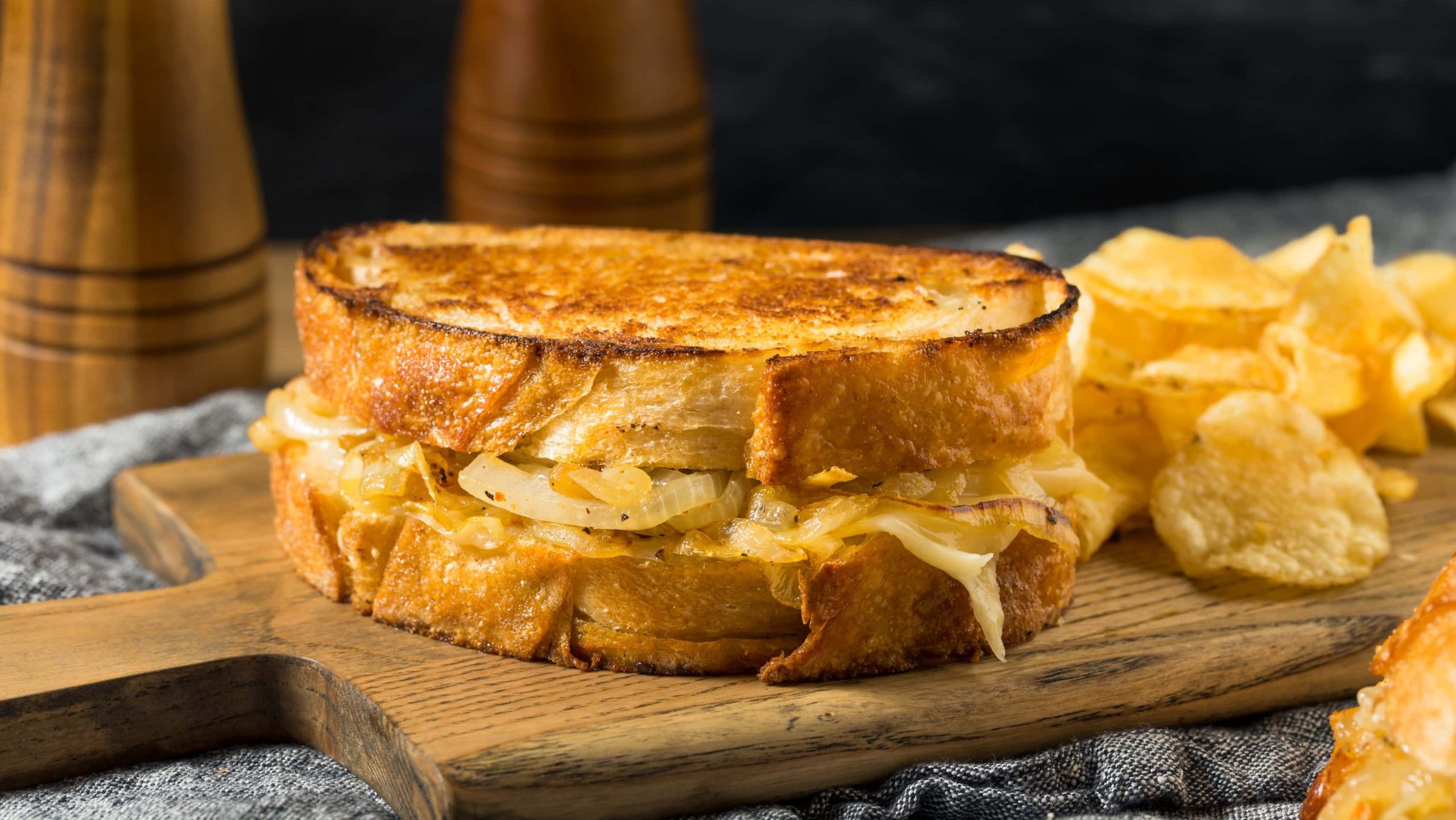 French Onion Cheese Sandwich mit Chips auf Holzbrett. Im Hintergrund Salzstreuer aus Holz. Frontalansicht.