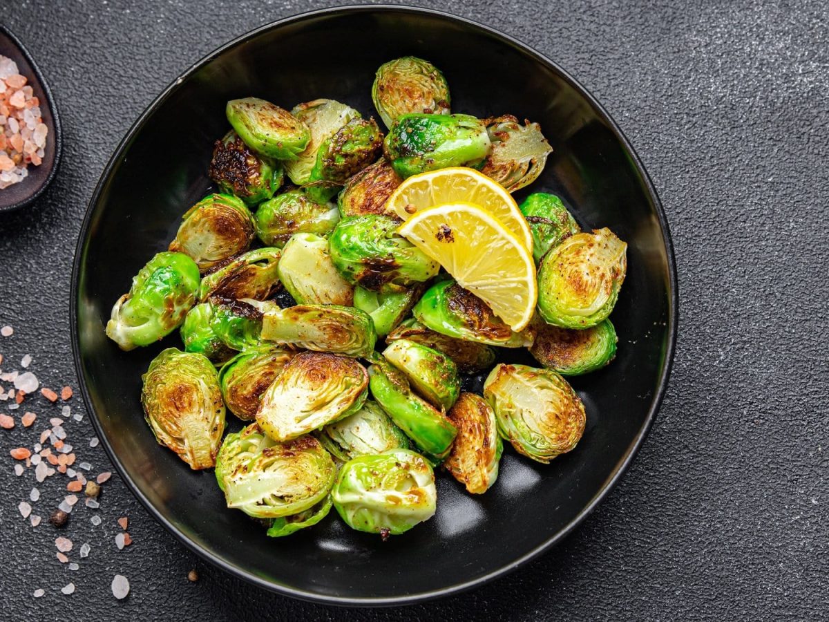 Eine Schale Rosenkohl mit Ahornsirup und Balsamico, darauf zwei Zitronenscheiben, daneben eine Schale grobes Salz, alles Draufsicht.