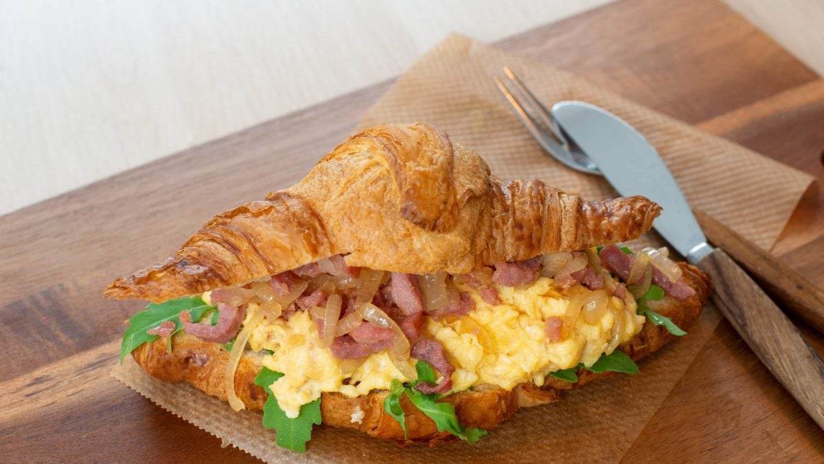 Gefülltes Croissant mit Rührei, Speckzwiebeln und Rucola auf Papier, Holzbrett und Holztisch. Im Bild Besteck mit Holzgriff. Draufsicht.