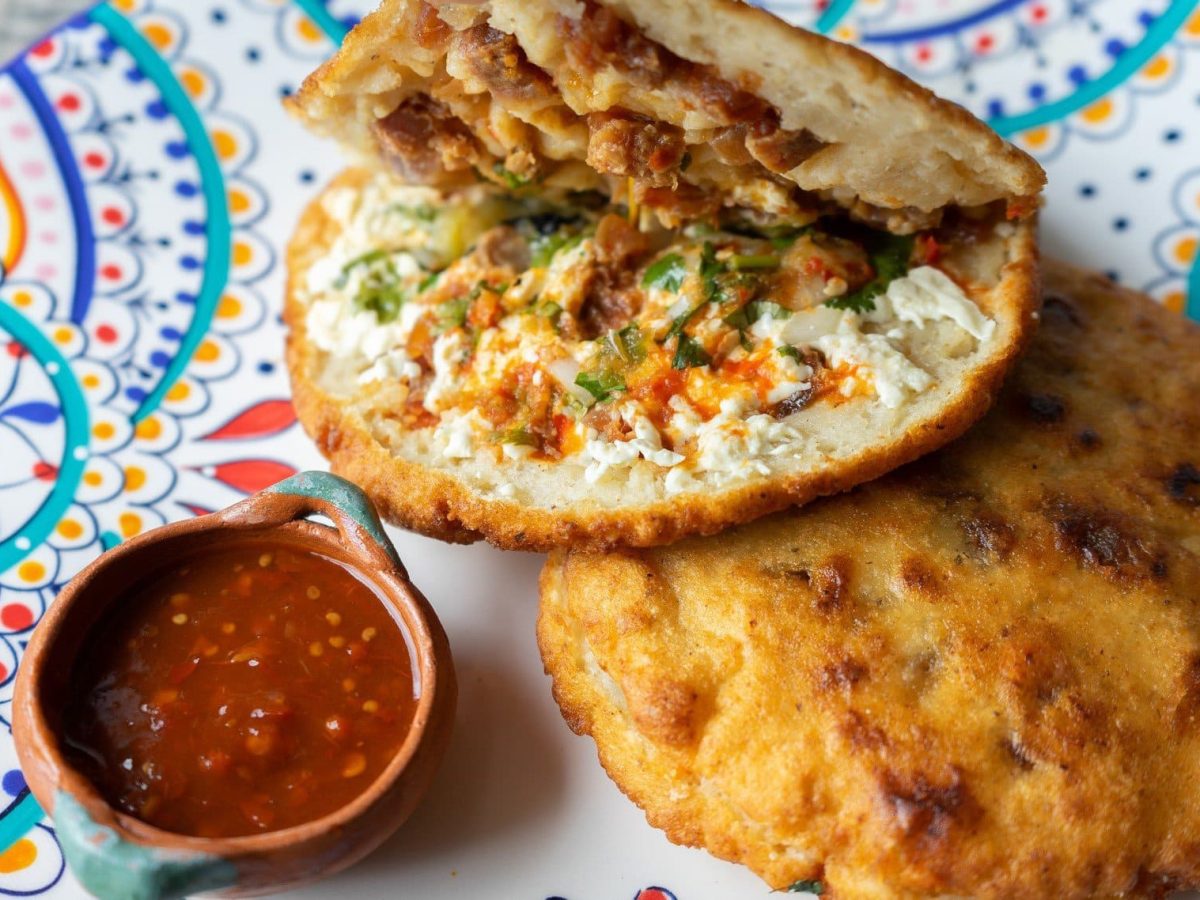 Zwei Gorditas de Chicharrón eines aufgeklappt mit einer Schale Salsa auf buntem Teller. Draufsicht.