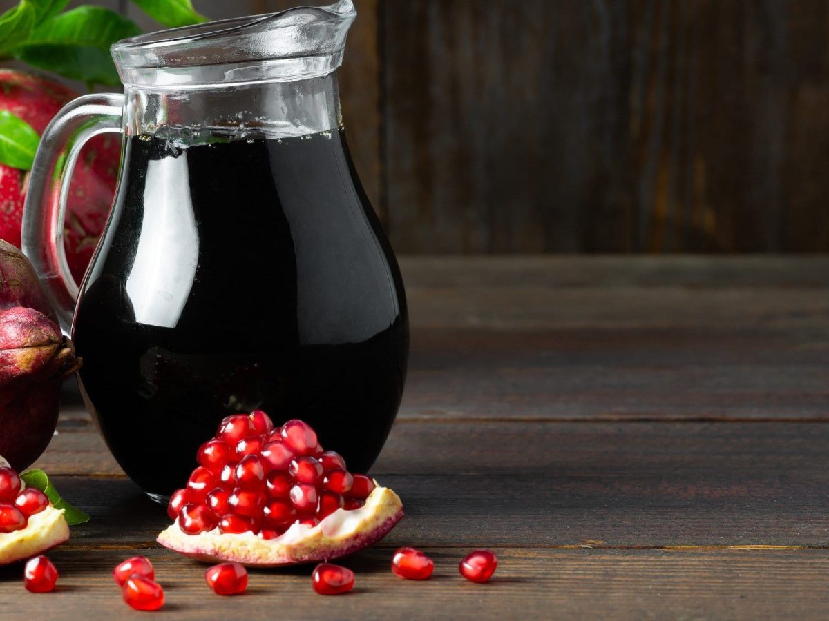 Selbst gemachte Granatapfel Melasse in Glaskaraffe auf Holz vor dunklem Hintergrund. Im Bild aufgeschnittener und ganzer Granatapfel mit Grün. Frontalansicht.