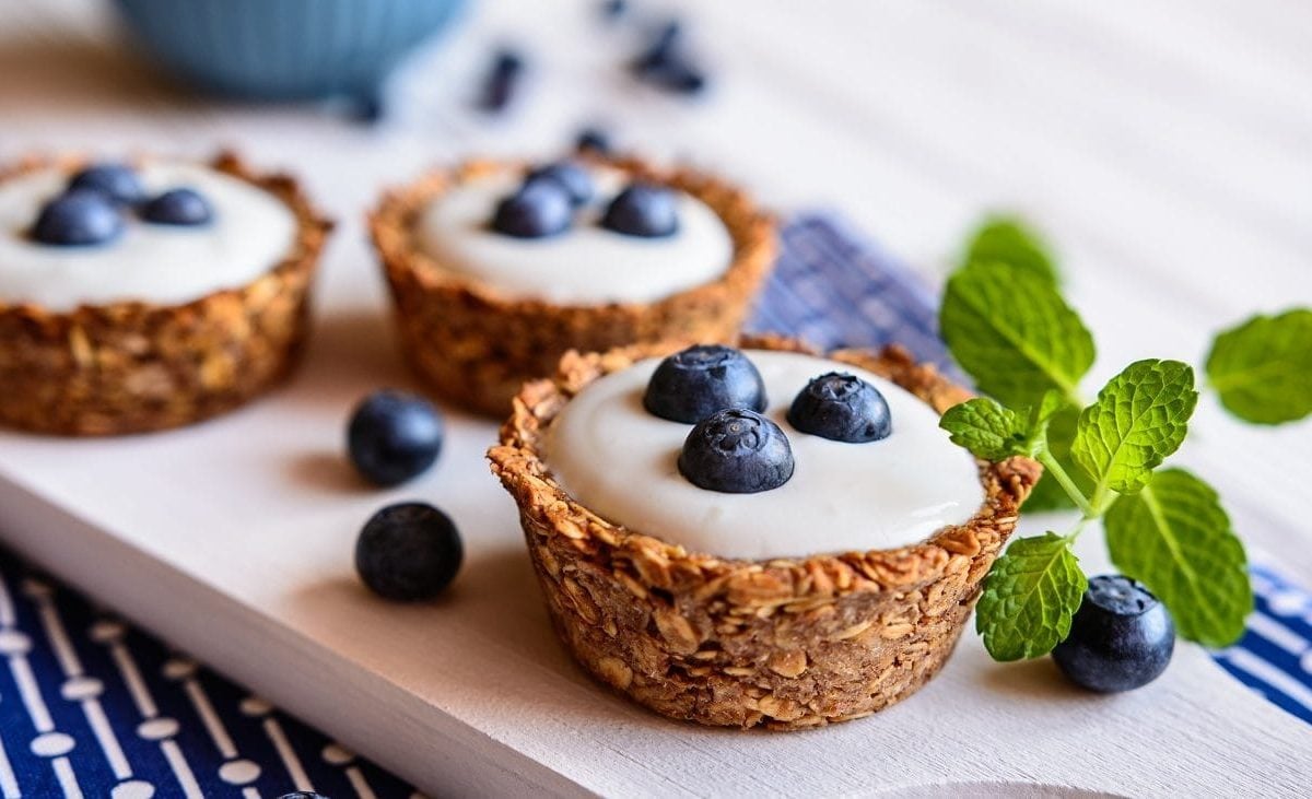 Frontalsicht: Auf einem hellen Untergrund liegt ein blaues, gemustertes Handtuch. Auf dem Handtuch liegt ein helles Holzbrettchen mit drei Granola-Cups mit Joghurt und heidelbeeren. Drumherum liegen frische Minzblätter.