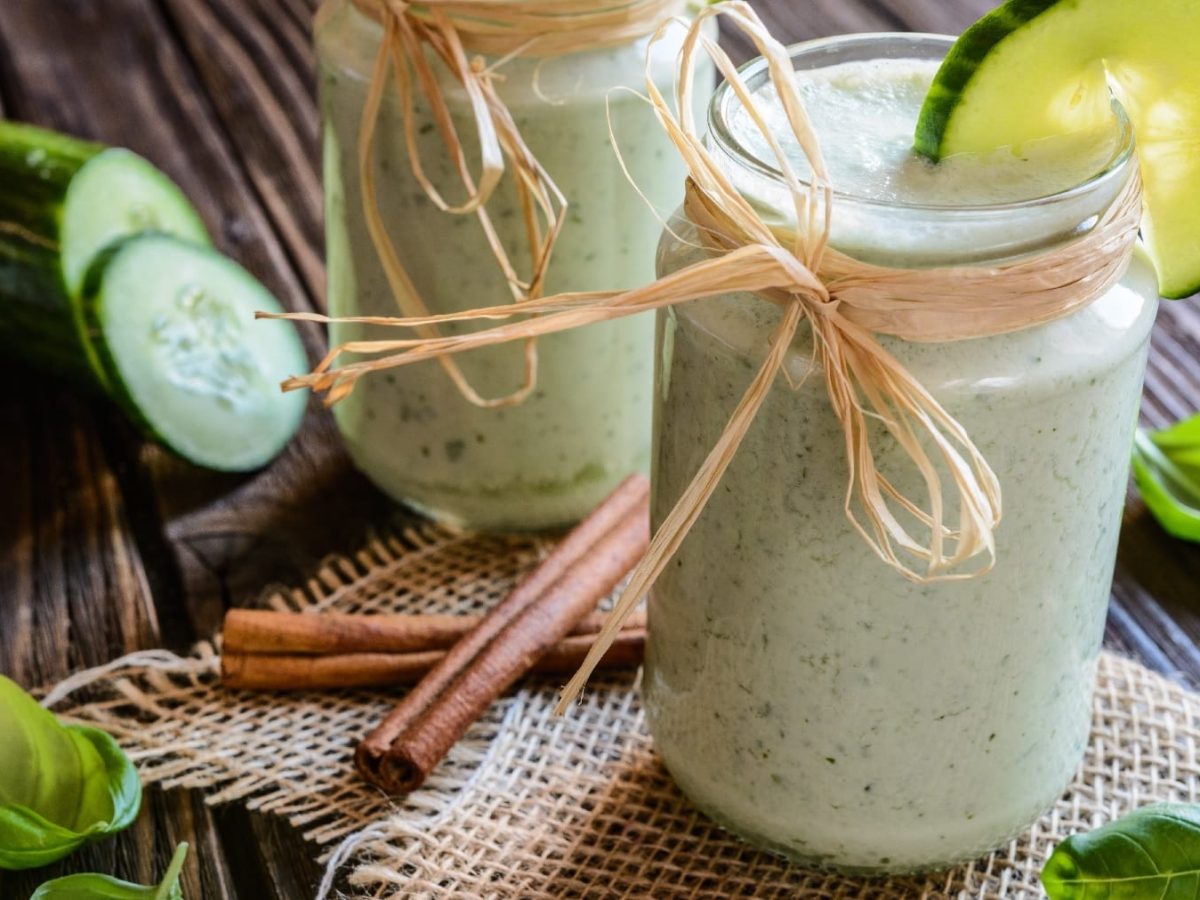 Zwei Gläser mit dem Gurken-Smoothie auf einem dunklen Holztisch. Daneben Gurke, Basilikum und Zimtstangen.