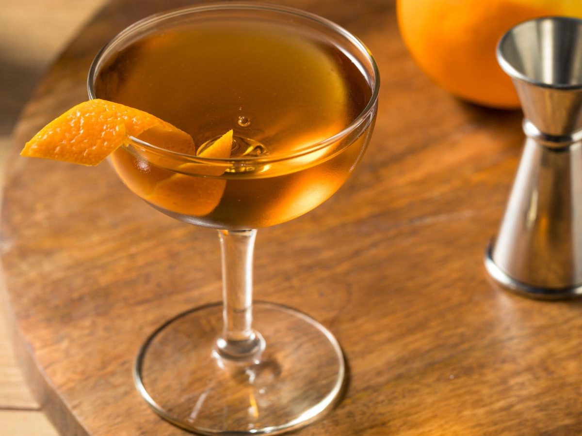 Hanky Panky Cocktail in Cocktailschale mit Orangenzeste auf Holztisch. Daneben Barmaß und eine Orange. Draufsicht.