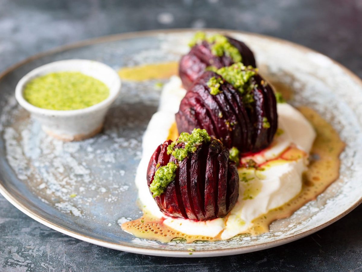 Drei gefächerte Hasselback-Betes auf einem Teller mit grünem Dressing und Jogurt.