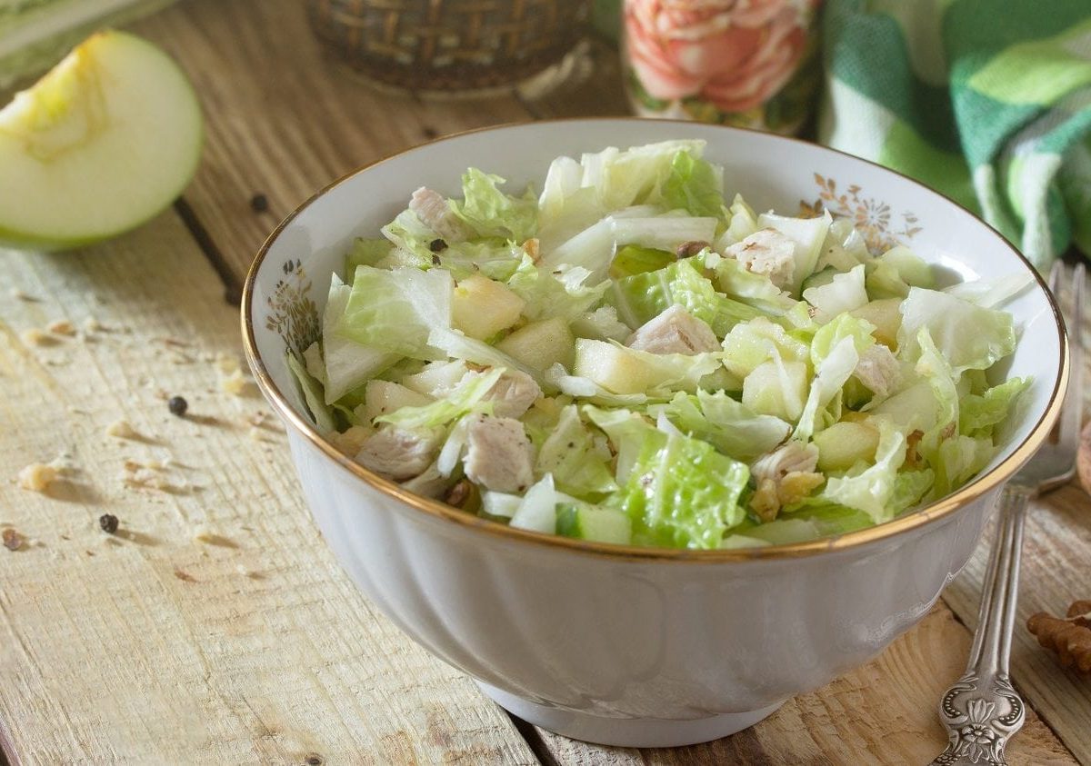 Schräge Draufsicht. Auf einem Untergrund aus Holz steht eine Porzellanschüssel mit einem Hühnchen-Apfel-Salat. Drum herum liegen Walnüsse, Gewürze sowie ein halber Apfel.