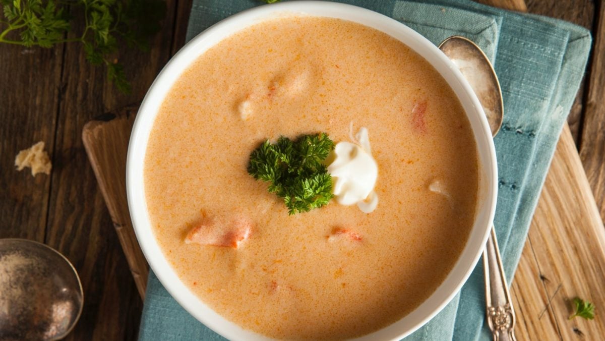 Draufsicht: Holztisch mit einer blauen Stoffservierte auf der eine weiße Porzellanschüssel steht. In der Schüssel eine Hummercremesuppe mit Cognac. Garniert mit etwas Crème fraîche und frischer Petersilie.