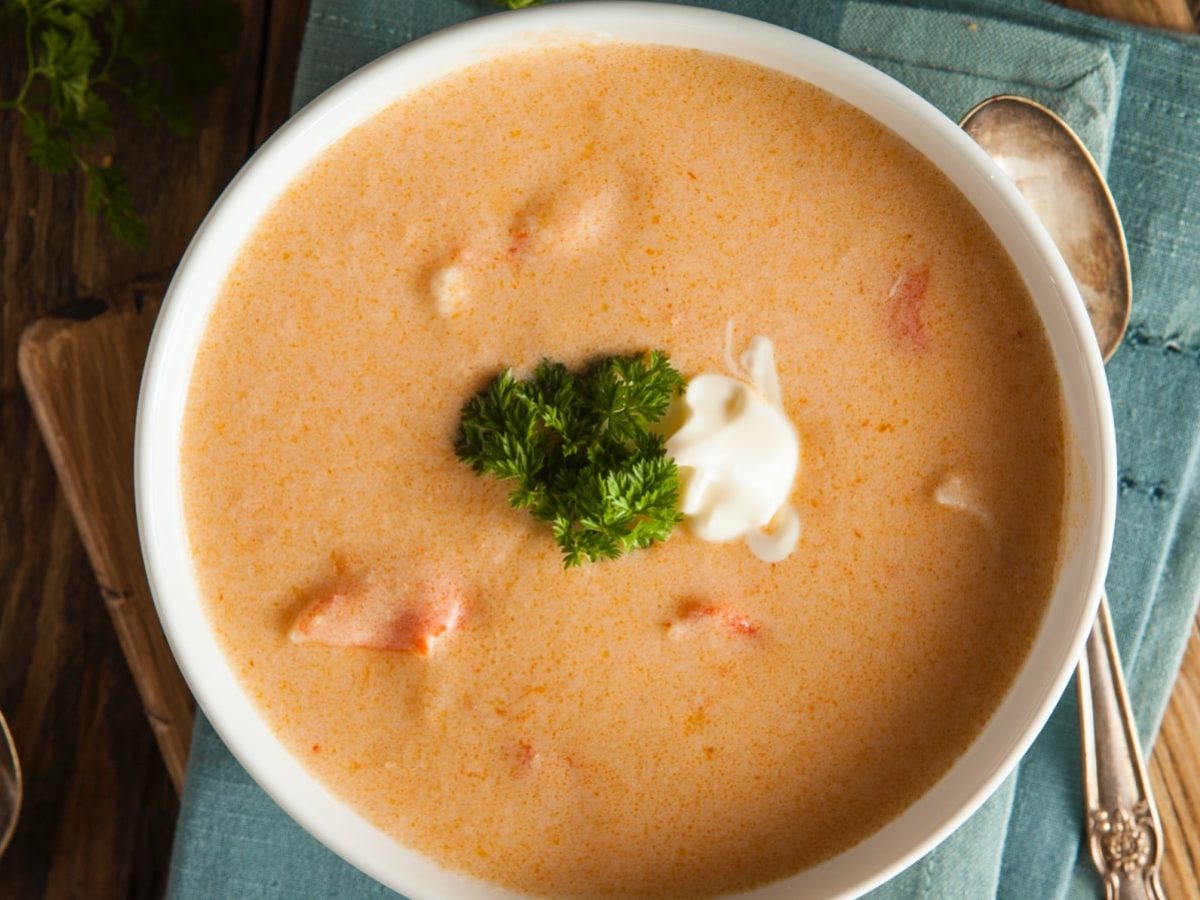 Draufsicht: Holztisch mit einer blauen Stoffservierte auf der eine weiße Porzellanschüssel steht. In der Schüssel eine Hummercremesuppe mit Cognac. Garniert mit etwas Crème fraîche und frischer Petersilie.