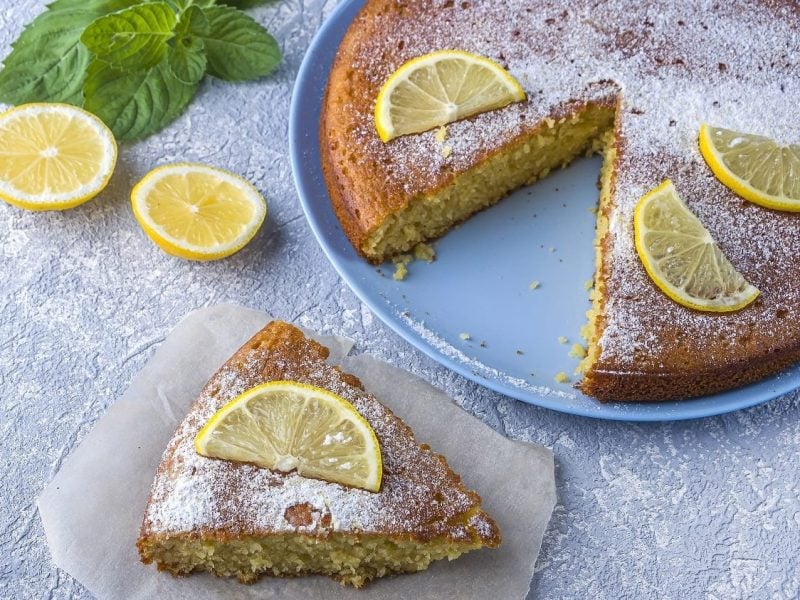 Vogelperspektive: Ein runder Grießkuchen mit Zitronen steht auf einer hellen Platte und diese auf einem hellen Untergrund. der Kuchen ist angeschnitten. Ein Stück liegt auf einem teller neben der Kuchenplatte.