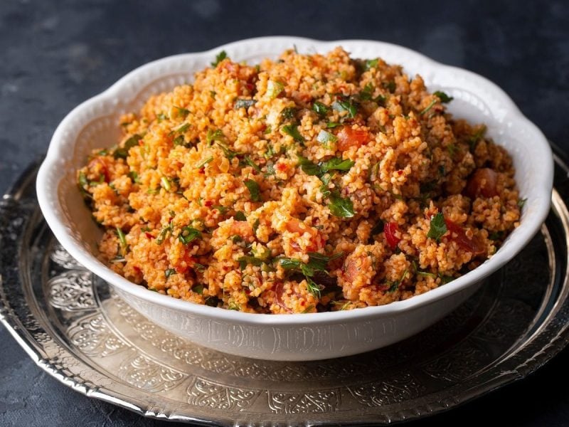 Frontalsicht. Auf einem dunklen Untergrund steht auf einem silberenen verzierten runden Teller eine eine weiße Porzellanschüssel mit dem bulgursalat kisir.