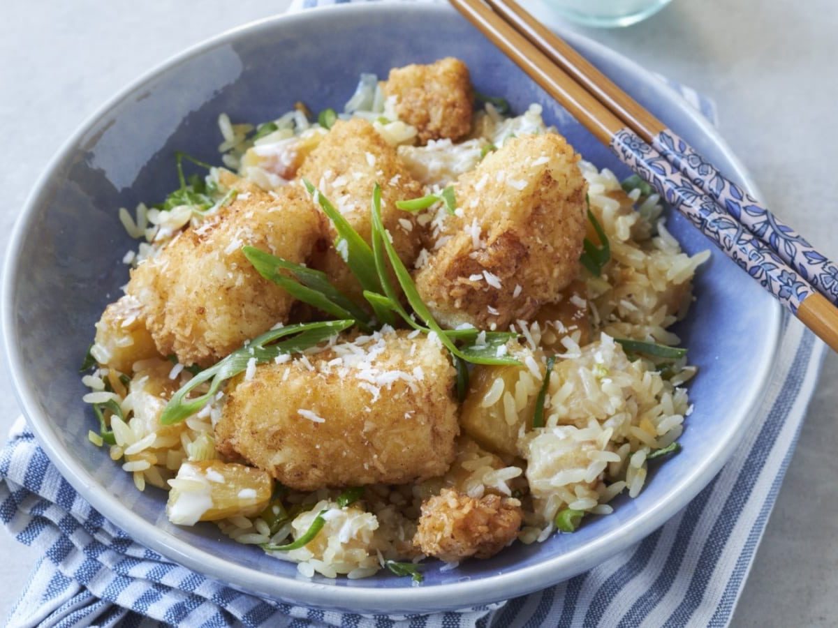 Draufsicht: Grauer Untergrund auf dem eine Schüssel auf einer blau-weißen Stoffservierte steht. In der Schüssel Kokos-Fisch-Nuggets mit Ananas-Reis.