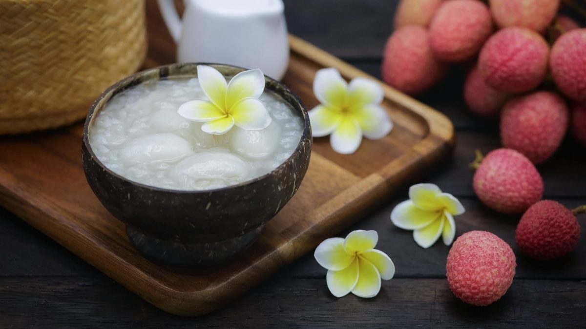 Eine braune Schale mit dem Litschi-Kokos-Pudding auf einem Holzbrett. Drum herum exotische Blüten, Litschis und ein Holzkorb.