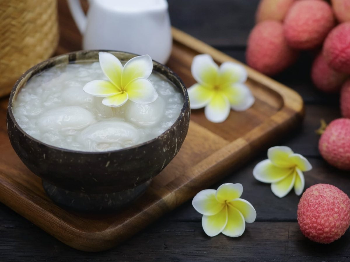 Eine braune Schale mit dem Litschi-Kokos-Pudding auf einem Holzbrett. Drum herum exotische Blüten, Litschis und ein Holzkorb.