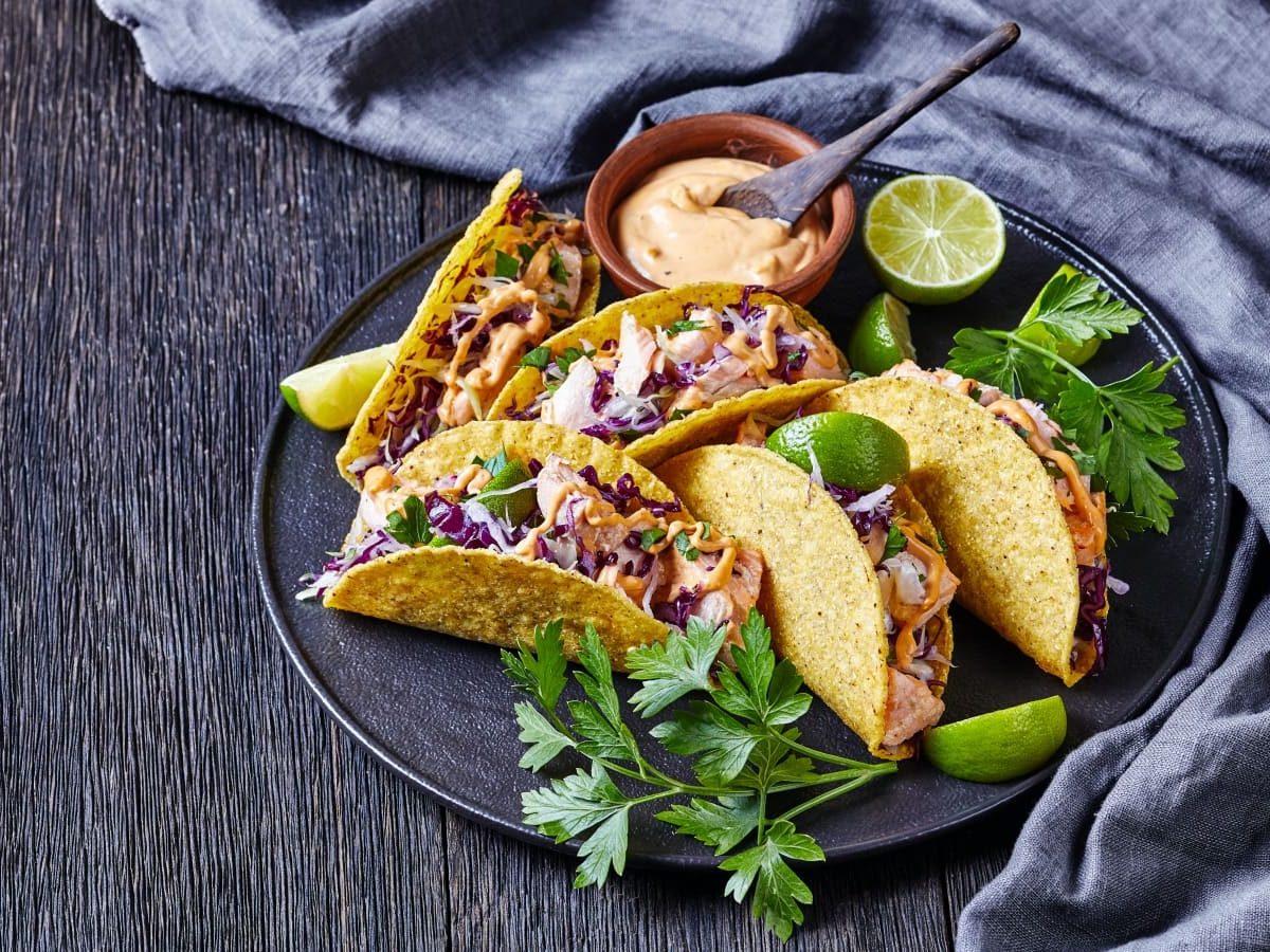 Draufsicht: Dunkler Untergrund mit einem grauen Geschirrtuch auf der rechten Seite. In der Bildmitte ein Teller mit fünf Lachs-Tacos mit Rotkohl und scharfer Joghurt-Sauce. Dazu frischer Koriander und Limette.