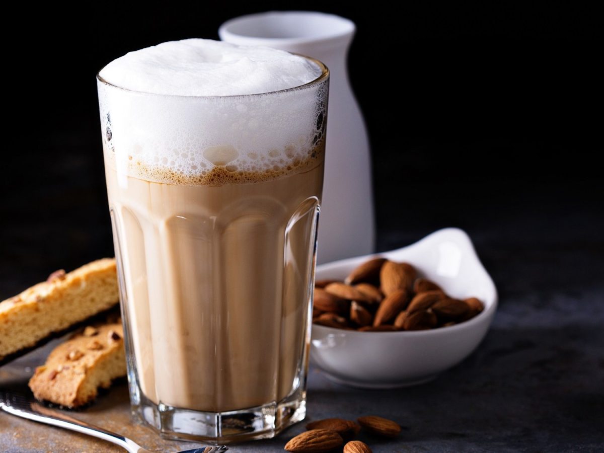 Ein Glas Mandel-Macchiato mit Milchschaum, daneben eine Schale Mandeln, ein Milchkännchen und Biscotti.