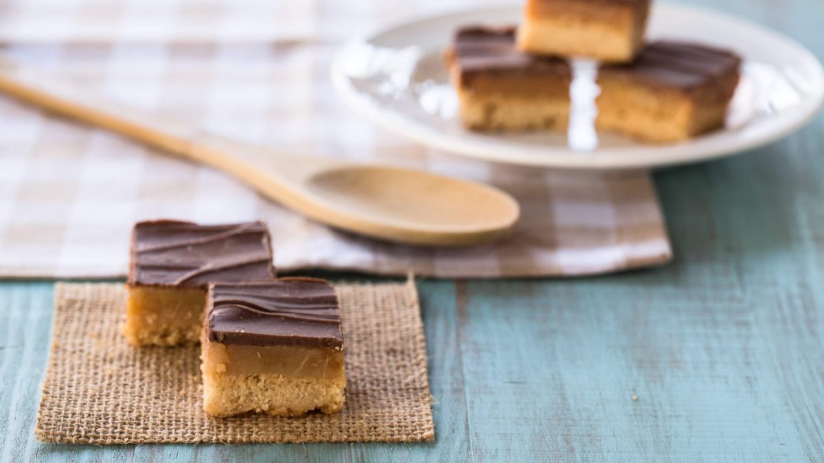 Millionaire's Shortbread in Stücken auf grauem Teller und Steinplatte. Im Hintergrund eine Schale mit Schokolade und Löffel. Frontalansicht.