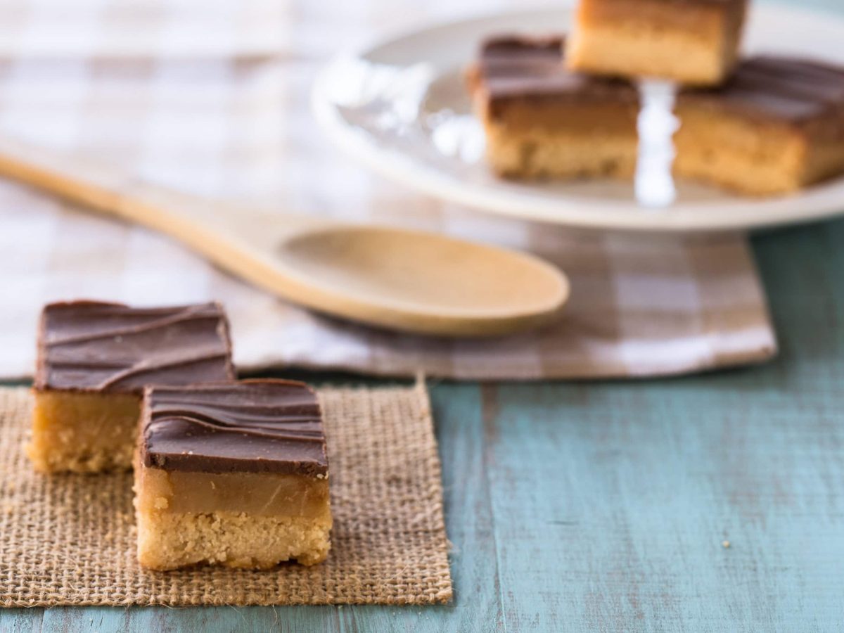 Millionaire's Shortbread in Stücken auf grauem Teller und Steinplatte. Im Hintergrund eine Schale mit Schokolade und Löffel. Frontalansicht.