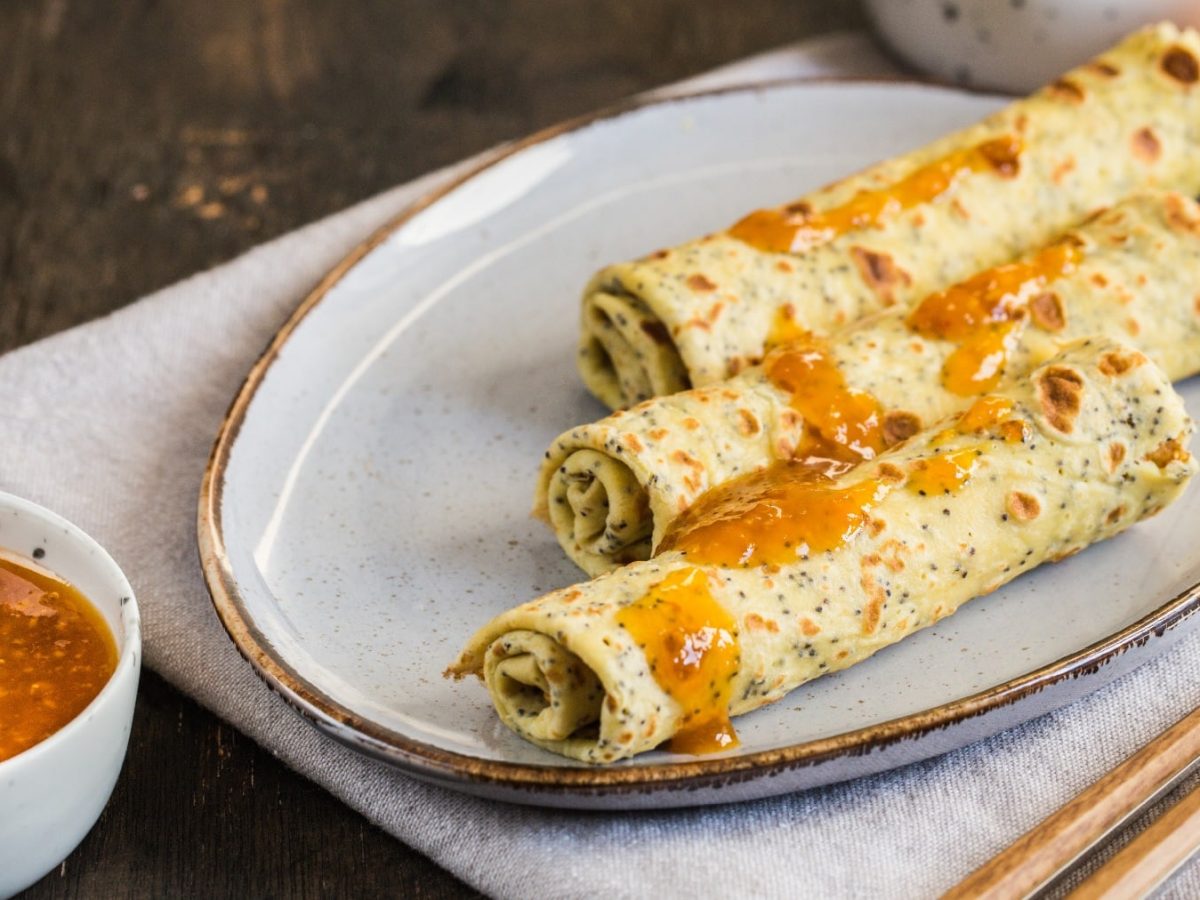 Ein weißer Teller mit den gerollten Mohn-Crepes auf dunklem Holztuntergrund. Daneben ein Schälchen mit Mandarinen-Marmelade.