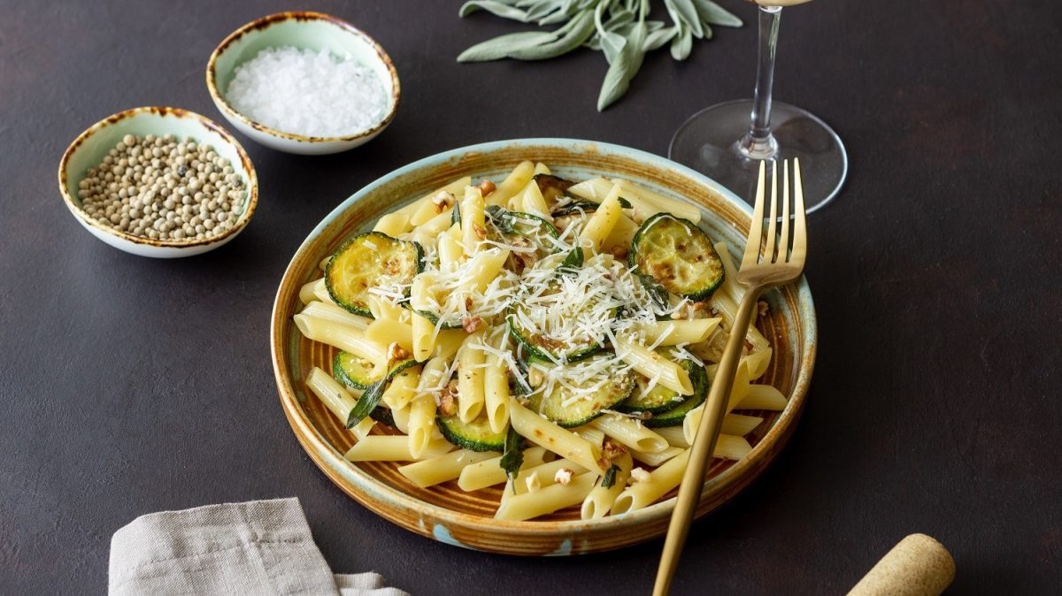 Schräge Draufsicht: Auf einem dunklen Untergrund steht ein Teller Penne mit Zucchini-Walnuss-Sauce. Auf dem Teller ist eine Gabel. Drum herum stehen Schälchen mit Gewürzen.