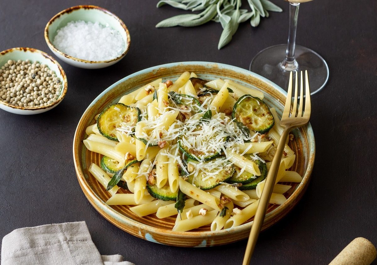 Schräge Draufsicht: Auf einem dunklen Untergrund steht ein Teller Penne mit Zucchini-Walnuss-Sauce. Auf dem Teller ist eine Gabel. Drum herum stehen Schälchen mit Gewürzen.