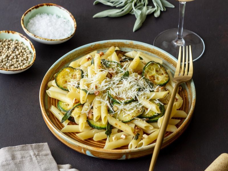 Schräge Draufsicht: Auf einem dunklen Untergrund steht ein Teller Penne mit Zucchini-Walnuss-Sauce. Auf dem Teller ist eine Gabel. Drum herum stehen Schälchen mit Gewürzen.