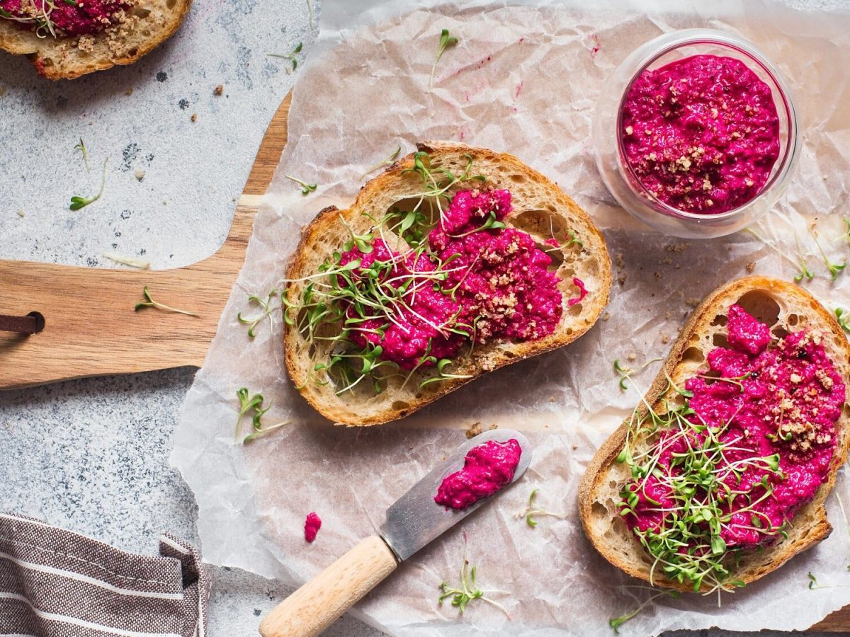 Brote mit Pink Hummus und Kresse auf einem Brett, daneben ein Glas Hummus und ein Messer, alles Draufsicht.