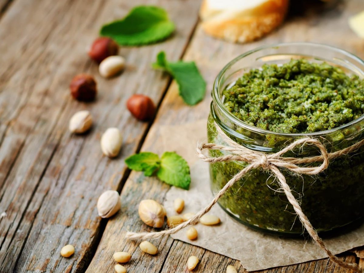 Ein Glas Pistazien-Pesto mit einer Schleife, daneben Pistazien und andere Nüsse.