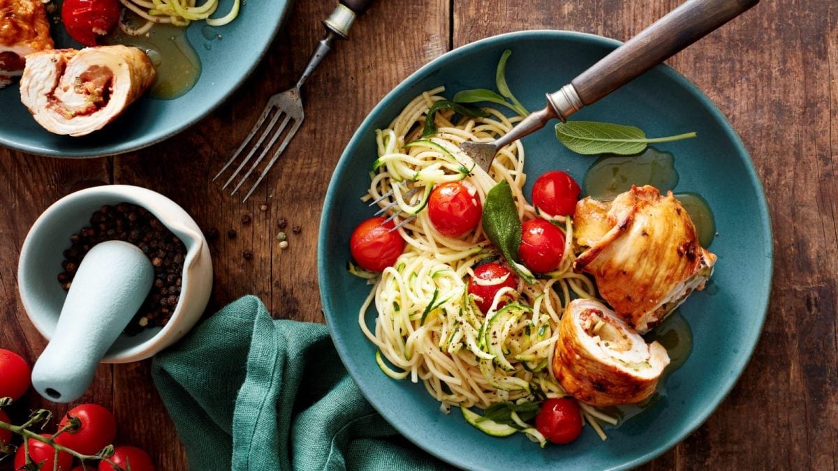 Draufsicht: Holzuntergrund mit einem türkisfarbenen Teller im Bildzentrum. Darauf zu sehen Puten-Zucchini-Rouladen mit Spaghetti, frischen Zucchini-Spiralen und Cherrytomaten. Außerdem auf dem Bild eine Stoffservierte und ein Mörser mit frischen Kräutern.