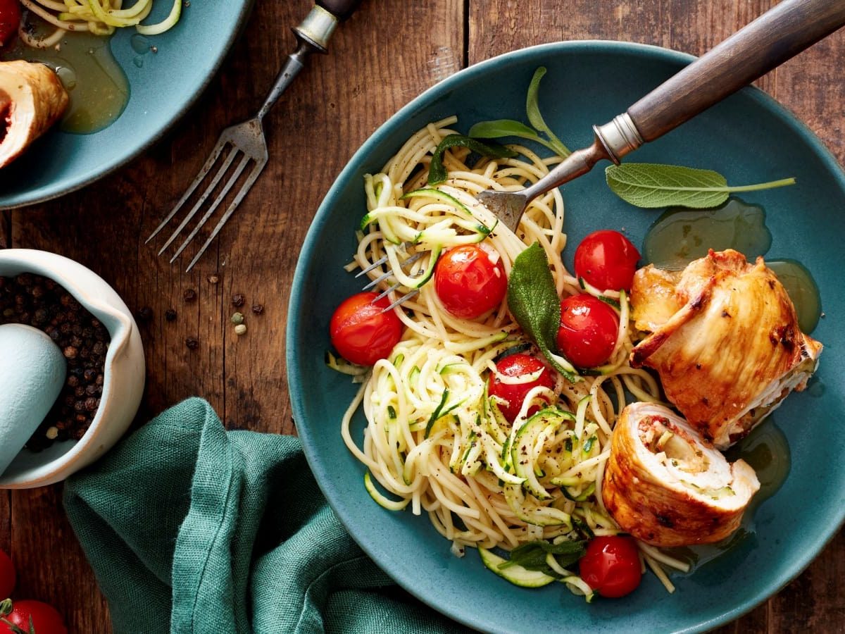 Draufsicht: Holzuntergrund mit einem türkisfarbenen Teller im Bildzentrum. Darauf zu sehen Puten-Zucchini-Rouladen mit Spaghetti, frischen Zucchini-Spiralen und Cherrytomaten. Außerdem auf dem Bild eine Stoffservierte und ein Mörser mit frischen Kräutern.