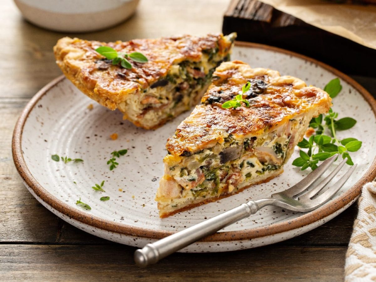 Ein weißer Teller mit braunem Rand und darauf ein Stück Quiche auf einem rustikalem Holztisch.