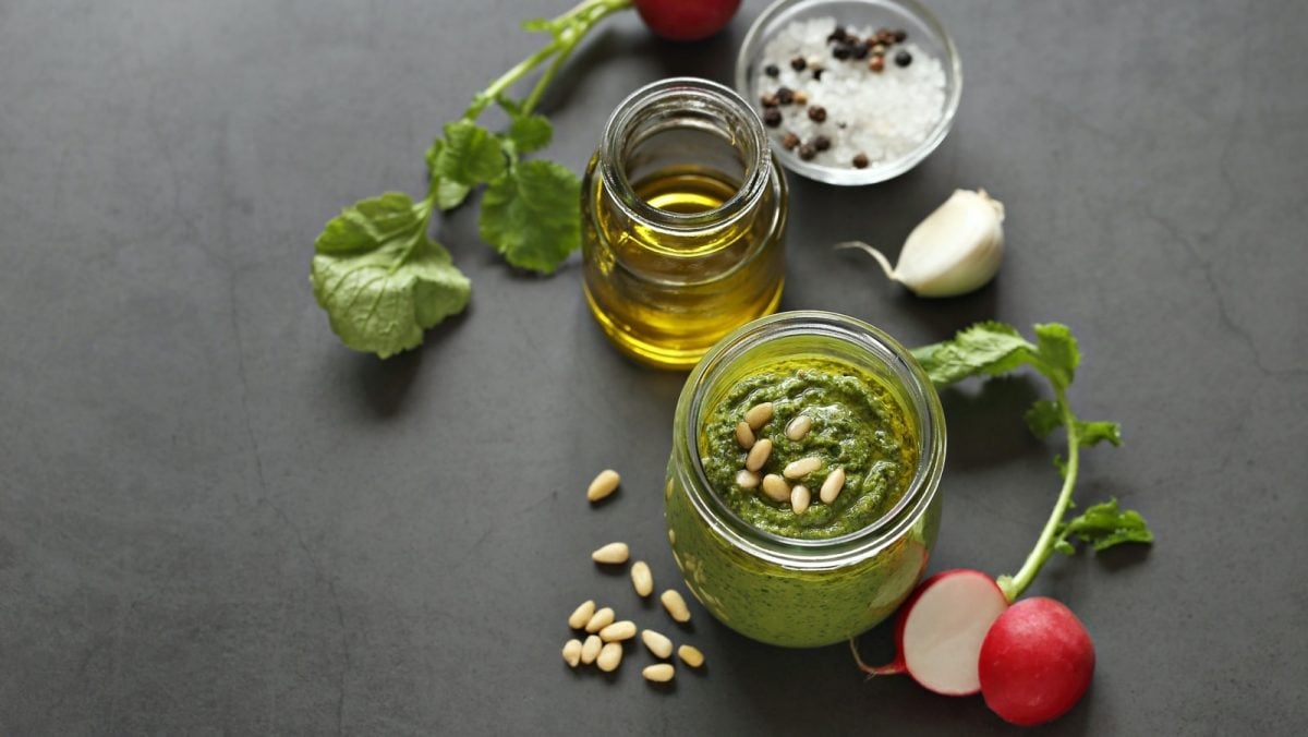 Draufsicht: Grauer Untergrund auf dem ein Schraubglas mit Radieschengrün-Pesto steht. Daneben ein weiteres Glas mit Olivenöl, einer frischen Knoblauchzehe, frischen Radieschen, Pinienkerne, grobkörniges Salz und Pfefferkörner.