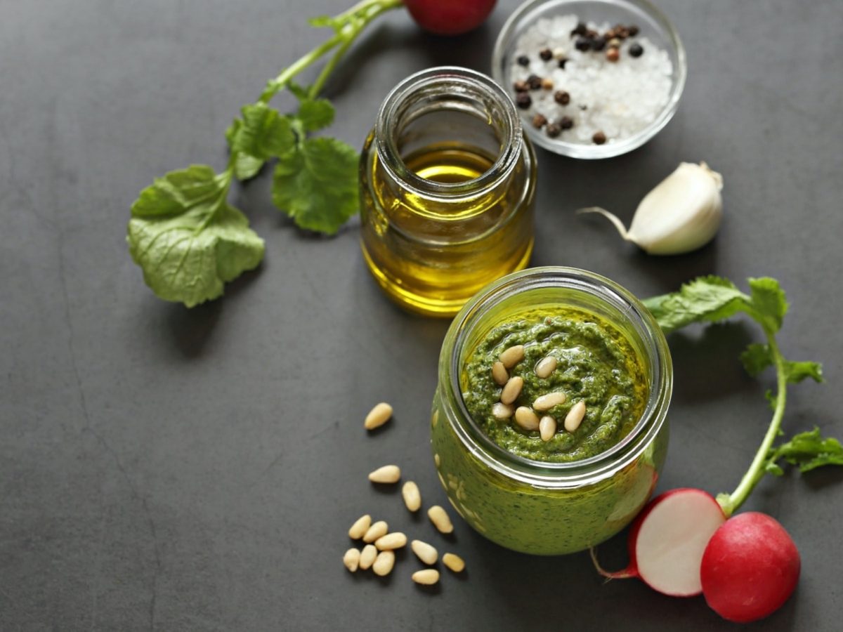 Draufsicht: Grauer Untergrund auf dem ein Schraubglas mit Radieschengrün-Pesto steht. Daneben ein weiteres Glas mit Olivenöl, einer frischen Knoblauchzehe, frischen Radieschen, Pinienkerne, grobkörniges Salz und Pfefferkörner.